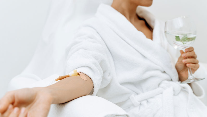 Close up of calm female patient sitting with tube and needle during IV infusion. Girl holding glass with lemon water and relaxing