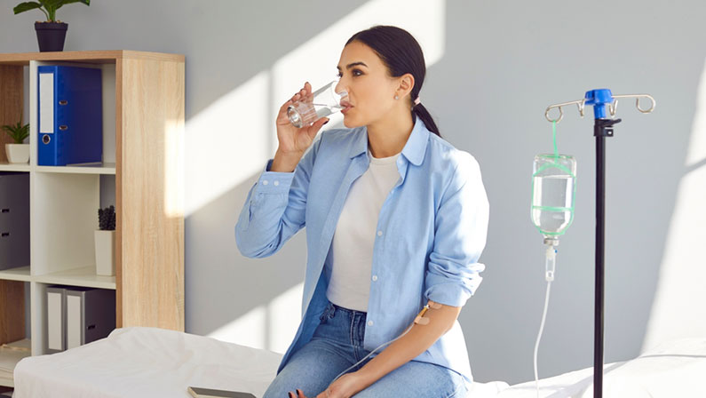 Young woman with sterile needle in arm sitting on medical bed by pole with IV drip bottle, drinking glass of water and receiving vitamin therapy infusion to boost immune system and suppress infection