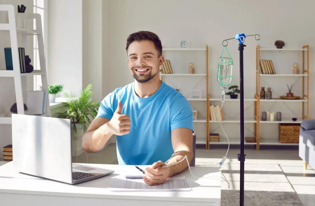 Happy smiling man sitting and working on a laptop showing thumb up sign while receiving IV drip