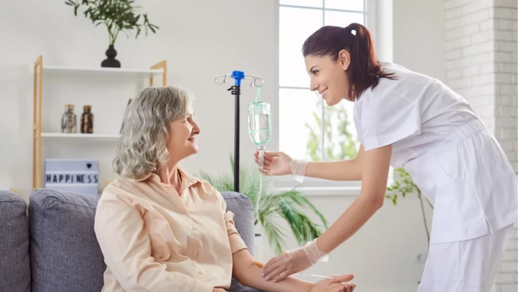 Young friendly nurse or caregiver help sick senior elderly woman patient in receiving IV drip infusion and vitamin injection therapy sitting on sofa in medical clinic or at home. Health care concept.