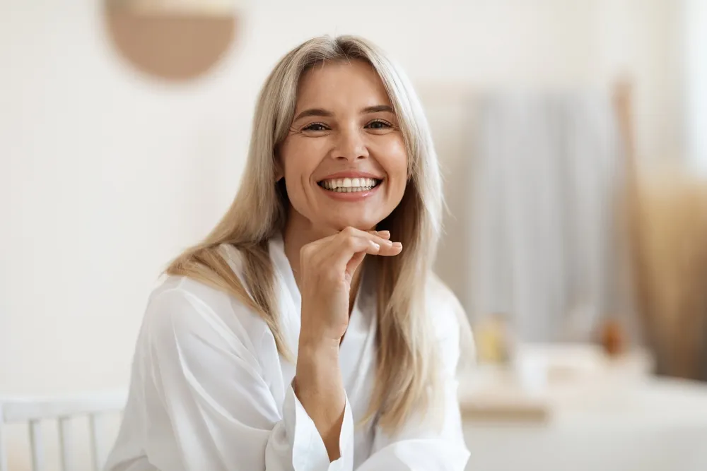 Happy attractive blonde woman in white silky bathrobe posing at white bathroom,